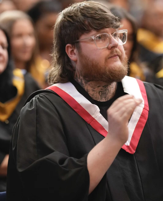 A male graduate with tattoos and glasses applauding during the graduation ceremony, wearing a black gown with a red and white stole. A male graduate with tattoos and glasses applauding during the graduation ceremony, wearing a black gown with a red and white stole.